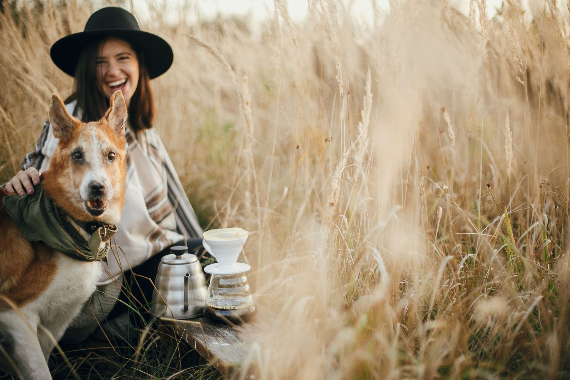 Stylish hipster traveler hugging her golden dog and smiling, brewing fresh coffee in rural countryside herbs in sunset light. Atmospheric rustic moment. Space for text. Traveling with pet